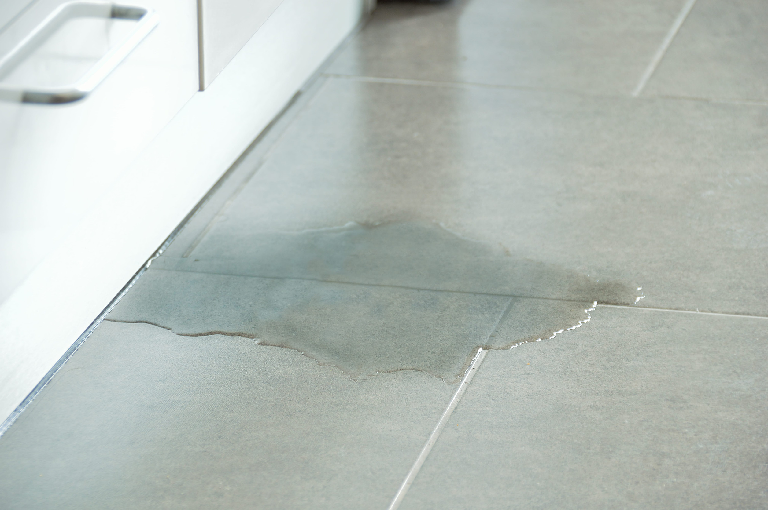 A pool of clear water spills across grey floor tiles next to the base of a white kitchen cabinet.

